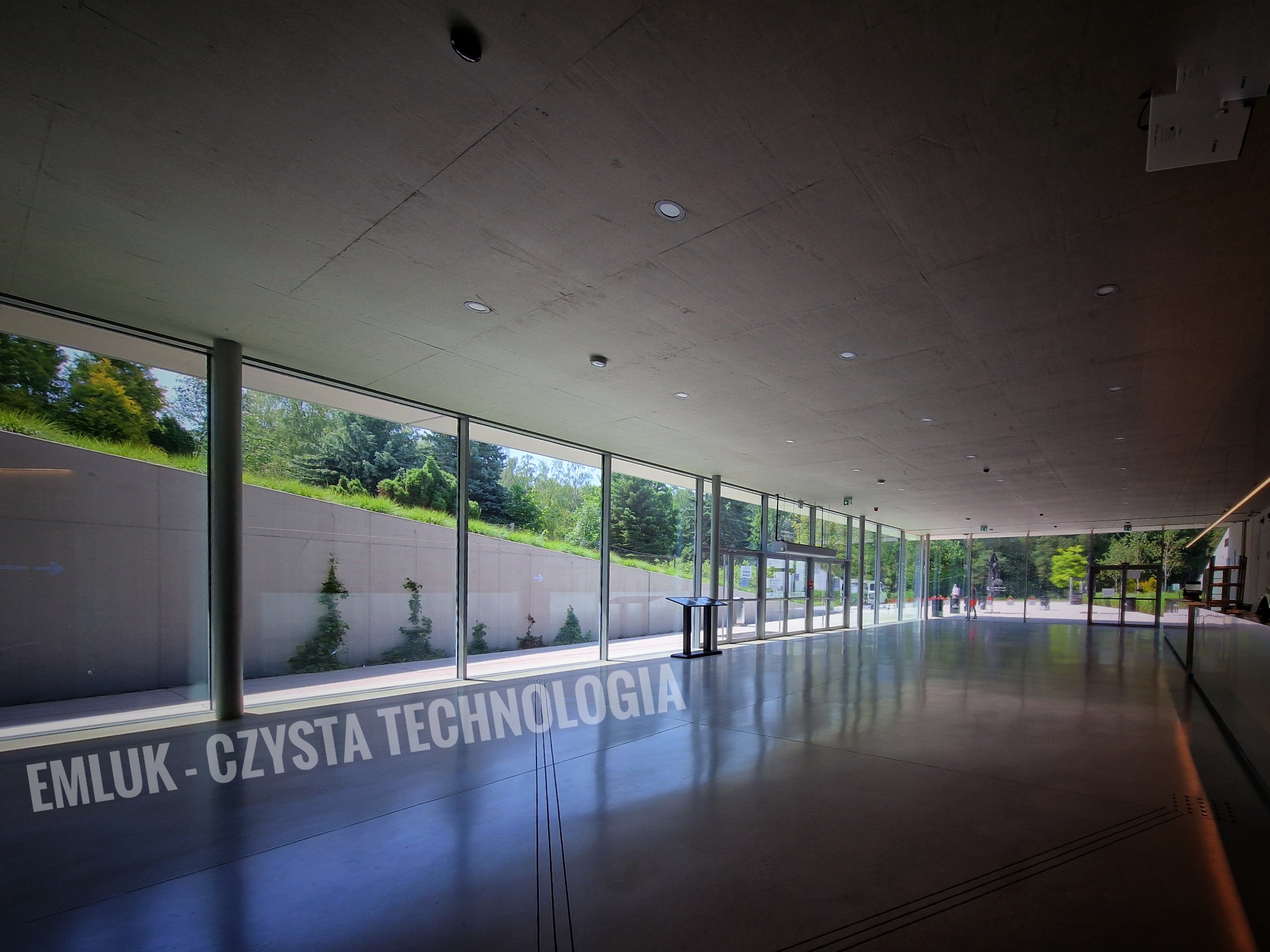 Wnętrze nowoczesnego budynku z przeszkloną ścianą, widok na zieleń na zewnątrz, na podłodze napis 'EMLUK - CZYSTA TECHNOLOGIA'.