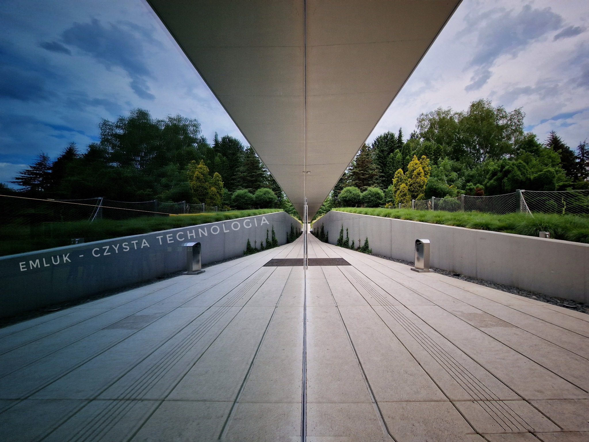 Architektoniczne ujęcie budynku z napisem 'EMLUK - CZYSTA TECHNOLOGIA', odbijającego się w szklanej fasadzie, ukazującego zieleń i niebo.