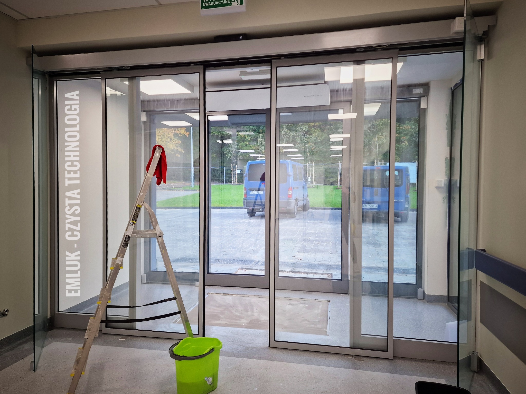 Aluminiowa drabina oparta o szklane, przesuwne drzwi z napisem EMLUK - CZYSTA TECHNOLOGIA, czerwona ścierka na szczeblu, obok zielone wiadro, widok na parking z dwoma niebieskimi busami.
