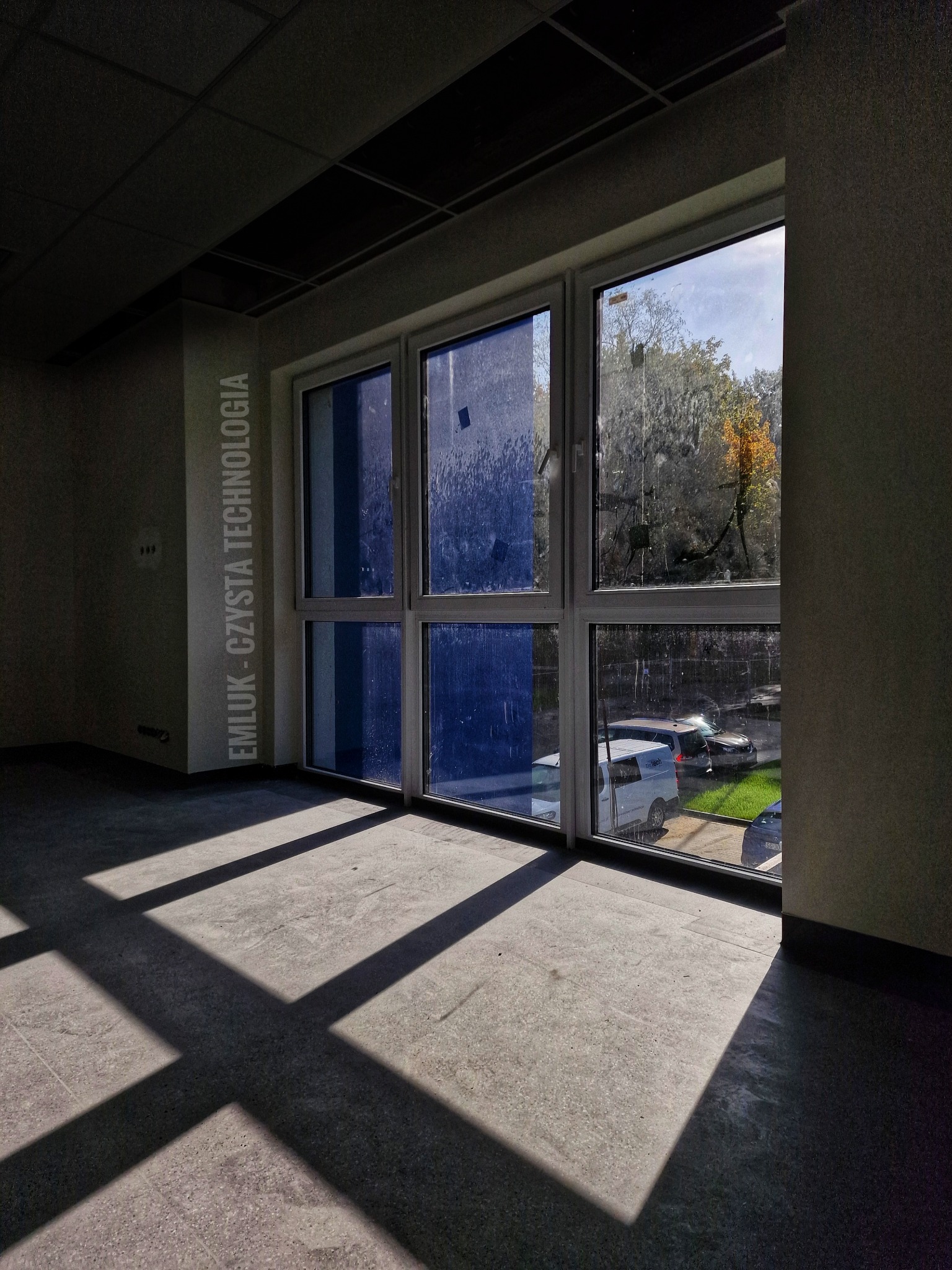 Wnętrze pokoju z dużym oknem, widoczne refleksy światła na podłodze, częściowo zaklejone szyby, widok na drzewa i parking na zewnątrz, napis 'EMLUK - CZYSTA TECHNOLOGIA' na ścianie.
