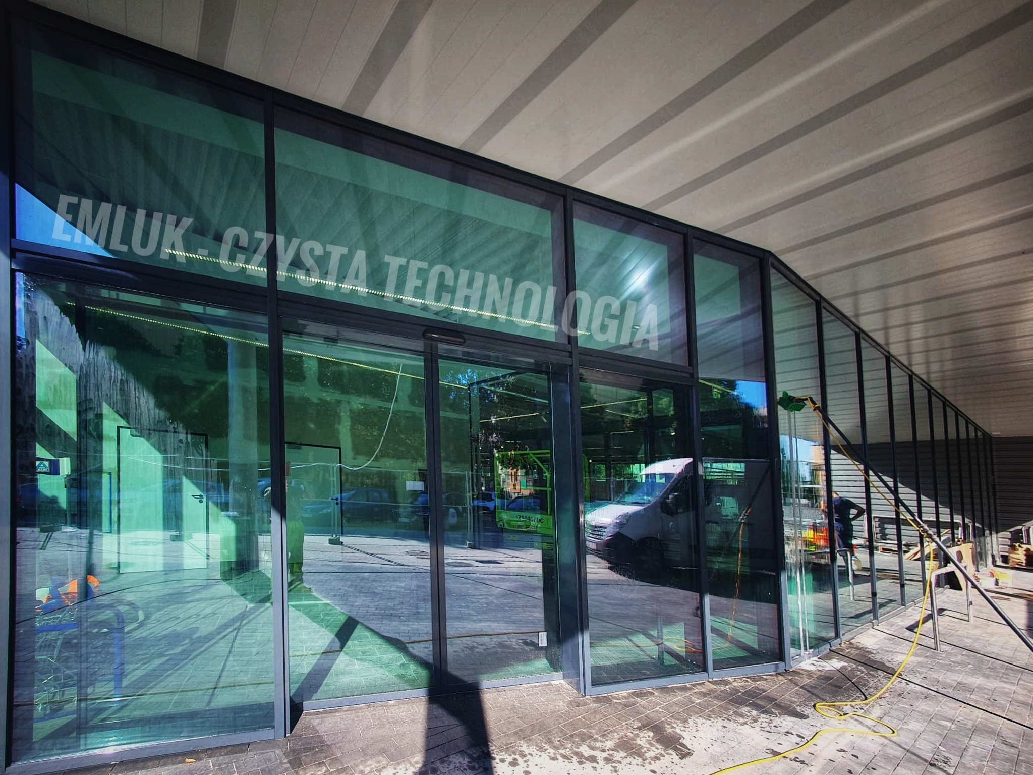 Mycie szklanej fasady budynku z napisem 'EMLUK - CZYSTA TECHNOLOGIA' za pomocą lancy teleskopowej i szczotki, widoczny samochód dostawczy i osoba w tle.