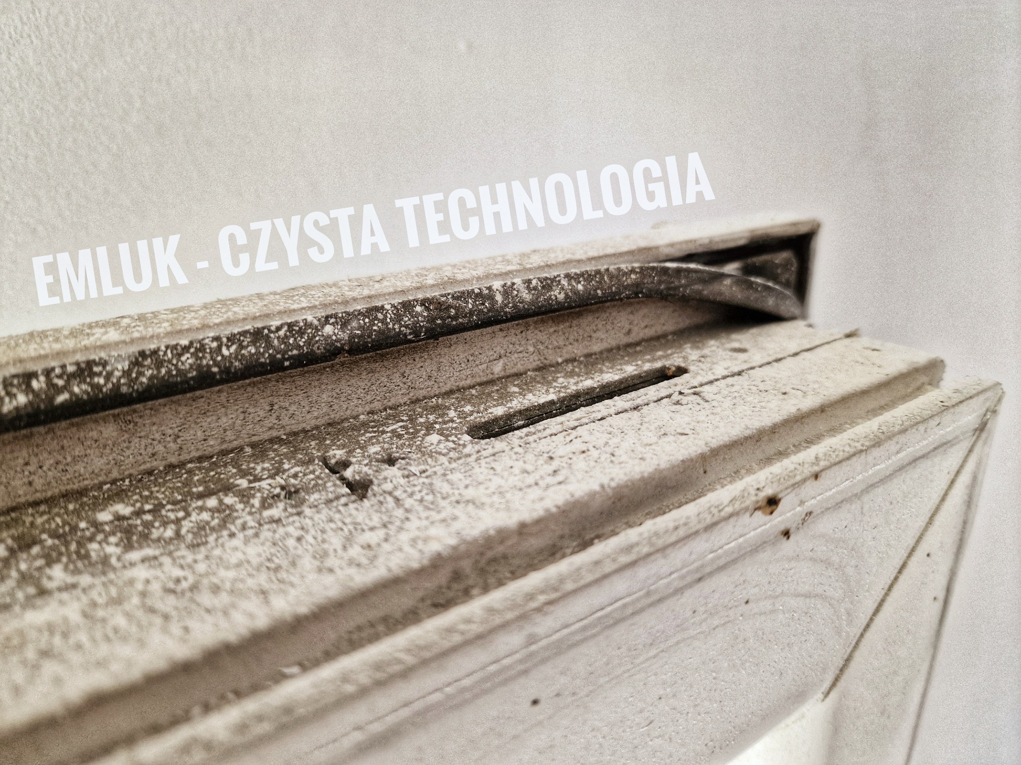 Bardzo brudne, zakurzone okno z widocznym logo 'EMLUK - CZYSTA TECHNOLOGIA' na górnej ramie.