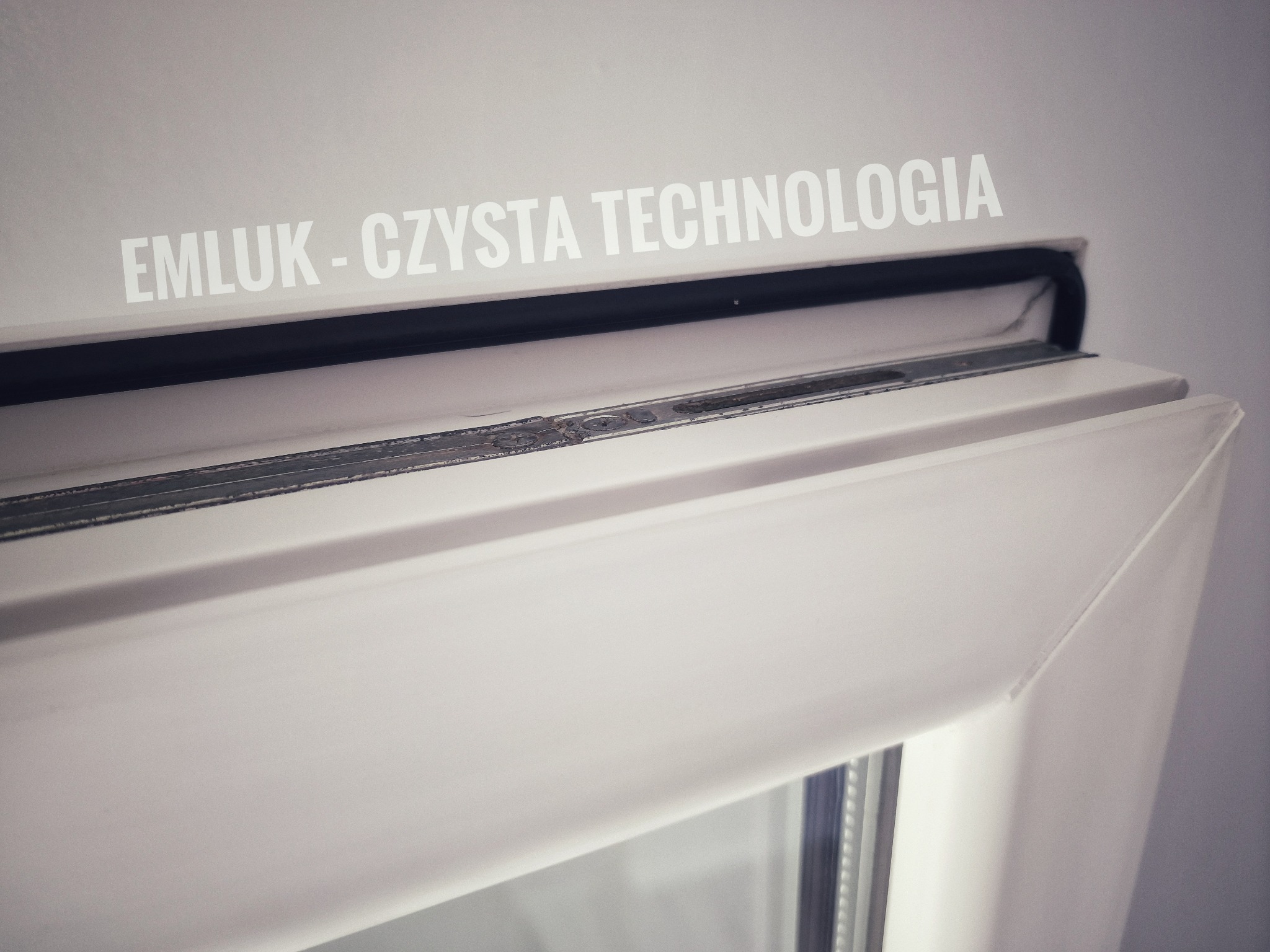 Detale ramy okiennej z widocznym uszczelnieniem i napisem 'EMLUK - CZYSTA TECHNOLOGIA' na ścianie.