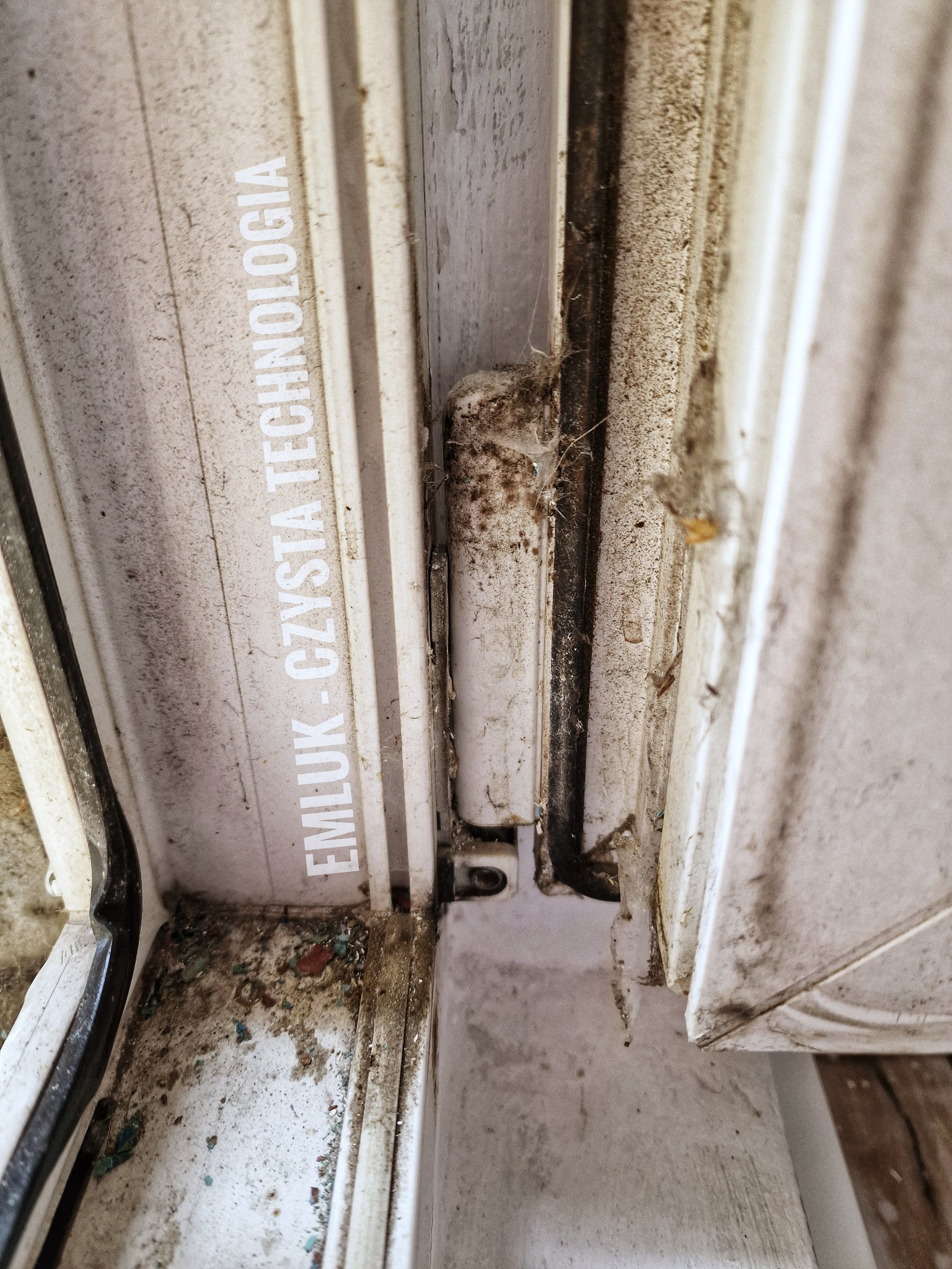 Silnie zabrudzony fragment ramy okiennej z widocznymi śladami kurzu, resztek roślinnych i pajęczyn, z napisem 'EMLUK - CZYSTA TECHNOLOGIA' na boku.