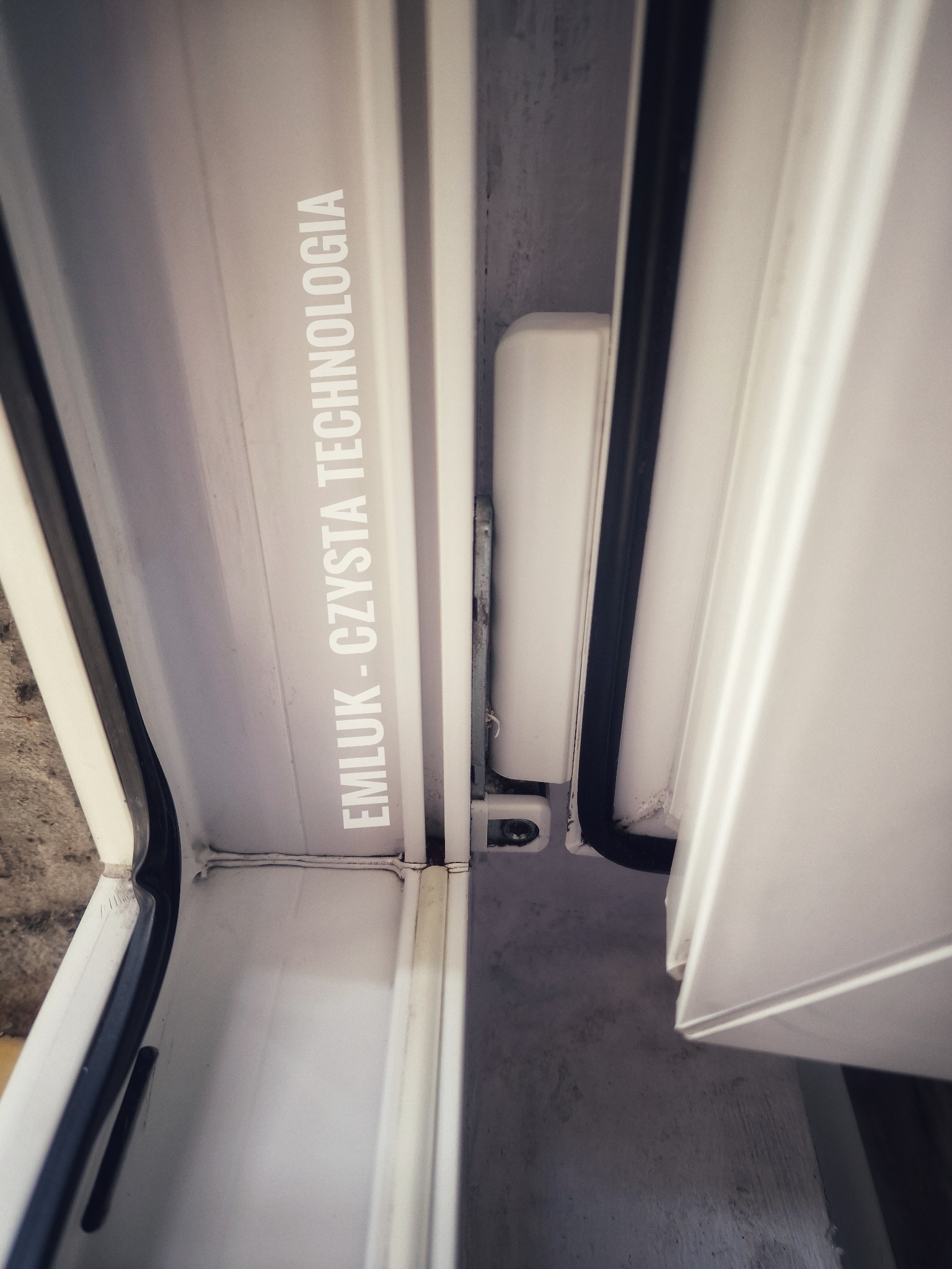 Fragment ramy okiennej z widocznymi uszczelkami i mechanizmem zamykającym, z nadrukowanym tekstem 'EMLUK - CZYSTA TECHNOLOGIA'.