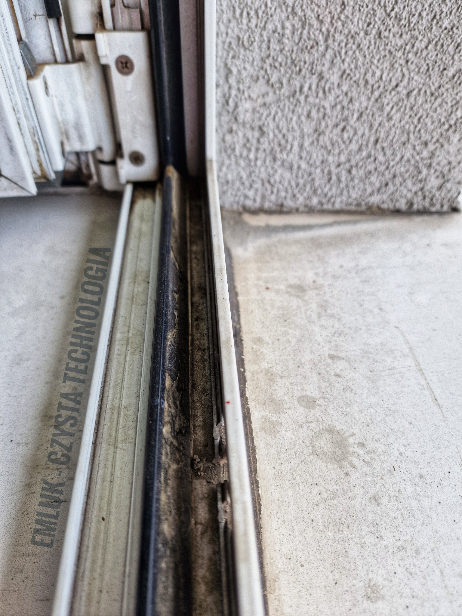 Brudna uszczelka okienna w ramie z widocznymi zabrudzeniami i kurzem wzdłuż krawędzi, fragment okna z widocznym logo firmy 'EMLUK-CZYSTA TECHNOLOGIA'.