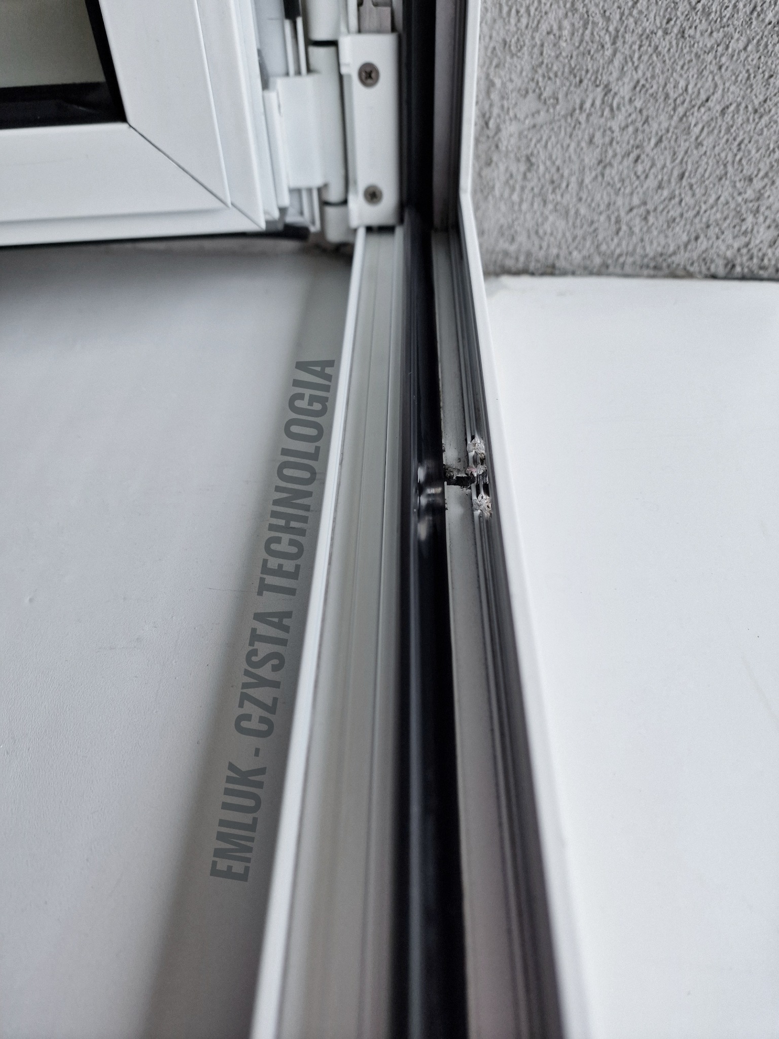 Szczegółowe ujęcie białej ramy okiennej PCV z widocznymi śladami brudu i uszczelkami, z fragmentem tekstu 'EMLUK - CZYSTA TECHNOLOGIA' w tle.