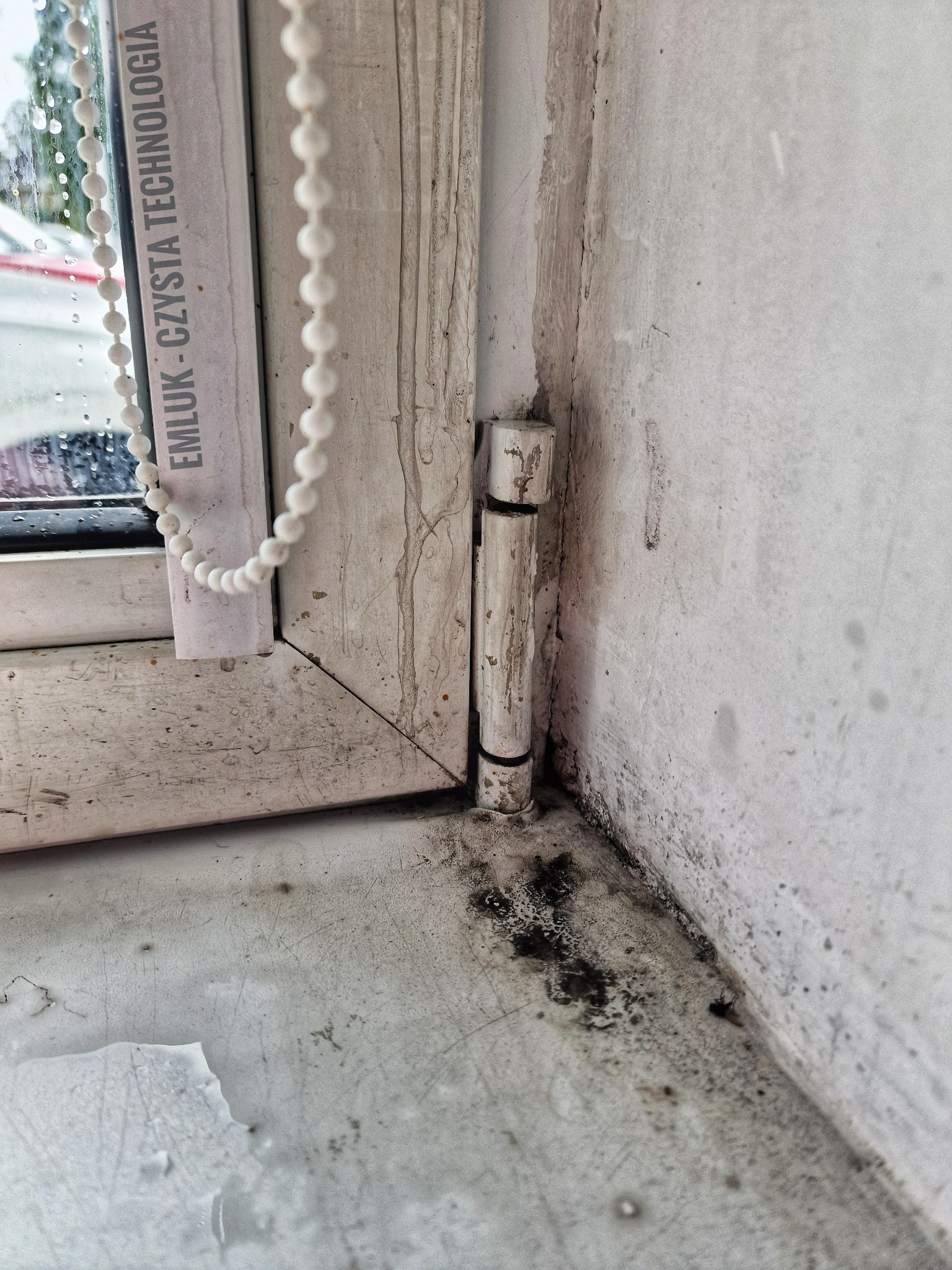 Brudne, białe okno PCV z widocznymi plamami pleśni w rogu i na parapecie, z elementami rolety łańcuszkowej i zawiasem.