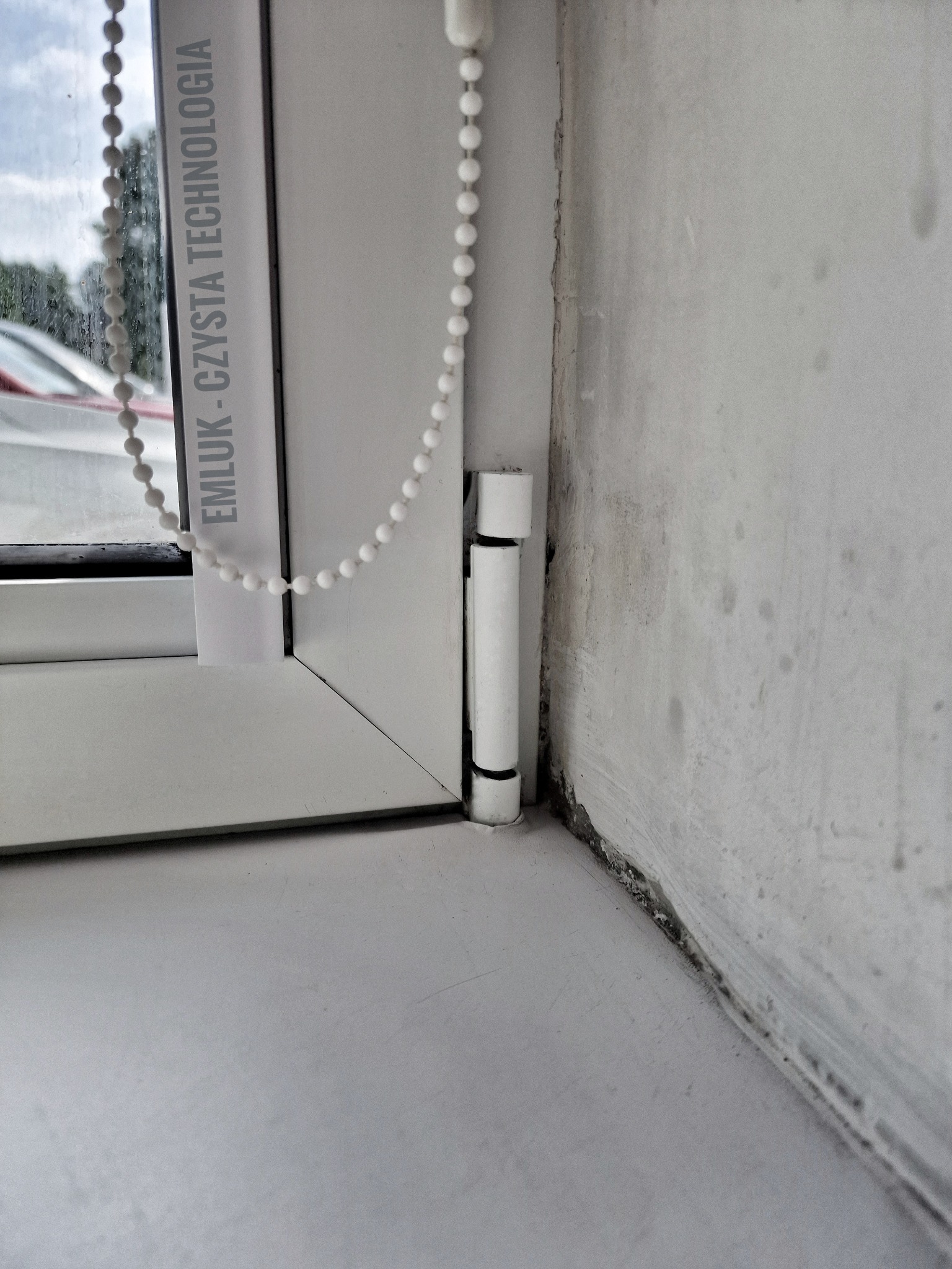 Fragment okna PCV z widocznym mechanizmem rolety i napisem 'EMLUK - CZYSTA TECHNOLOGIA', brudna ściana w tle.
