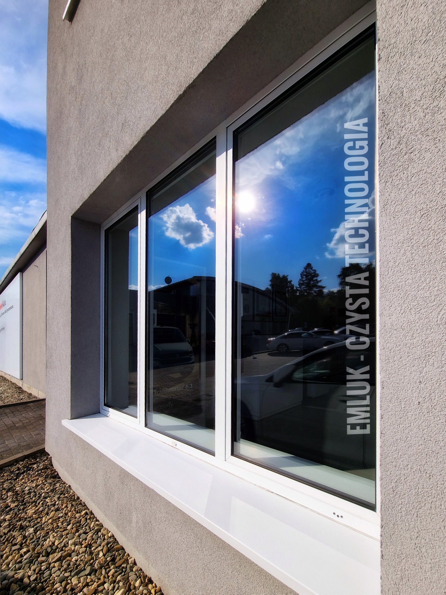 Trzyczęściowe okno z białymi ramami w szarej ścianie budynku, w szybach odbija się błękitne niebo z chmurami i budynki, na szybie pionowy napis 'EMLUK - CZYSTA TECHNOLOGIA'.