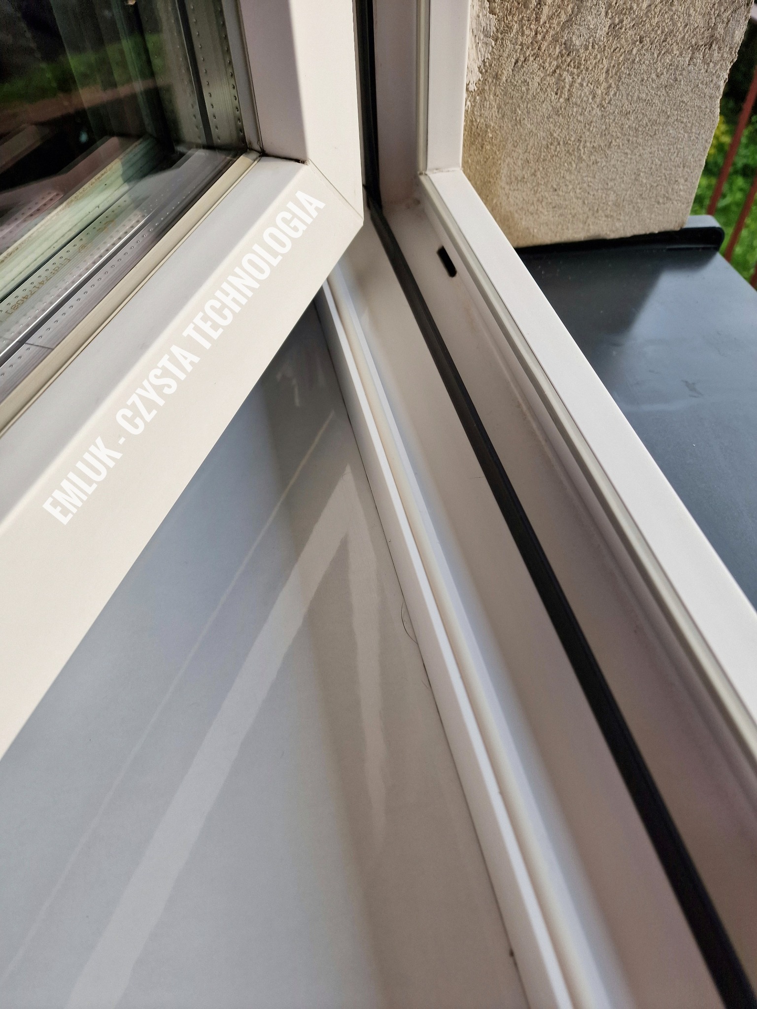 Wnętrze czystego, białego okna PCV z widocznym uszczelnieniem, z zewnątrz widoczny fragment muru i zieleń, na ramie okiennej nadrukowane hasło 'EMLUK - CZYSTA TECHNOLOGIA'.