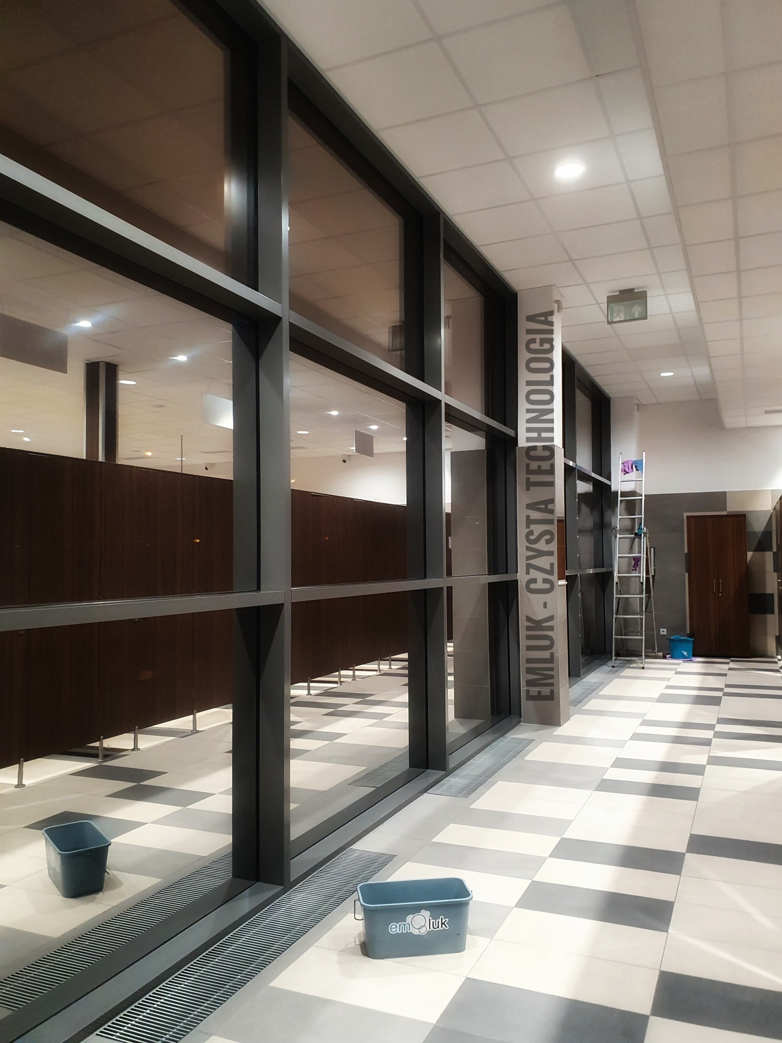 Duże okna w hali, widoczne wiadro z logo firmy sprzątającej, drabina z szmatkami i czyszczone okna w tle.