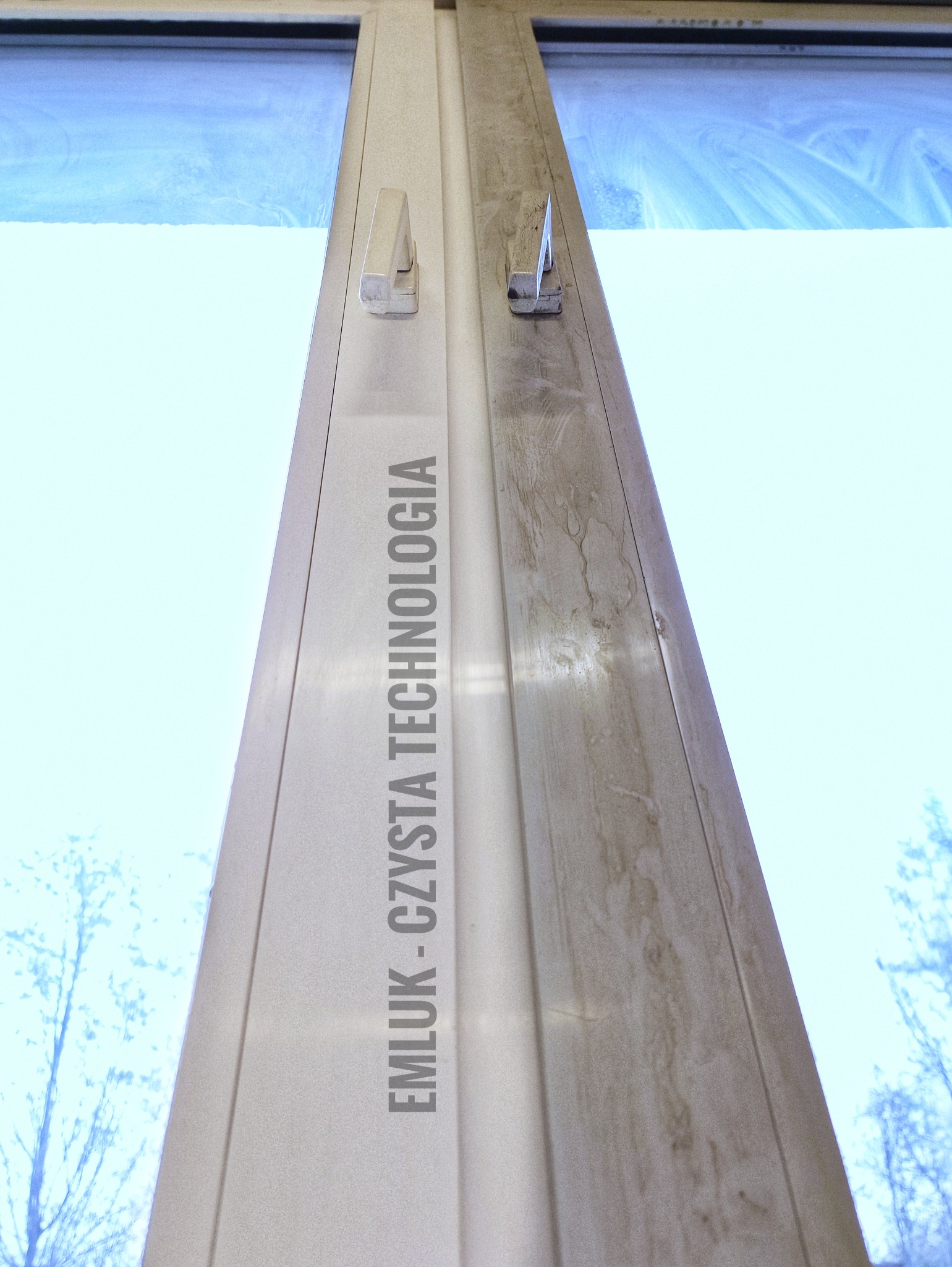 Okno PCV z widoczną różnicą czystości: prawa strona brudna, lewa świeżo umyta, z logo firmy 'EMLUK - CZYSTA TECHNOLOGIA' na ramie okiennej.