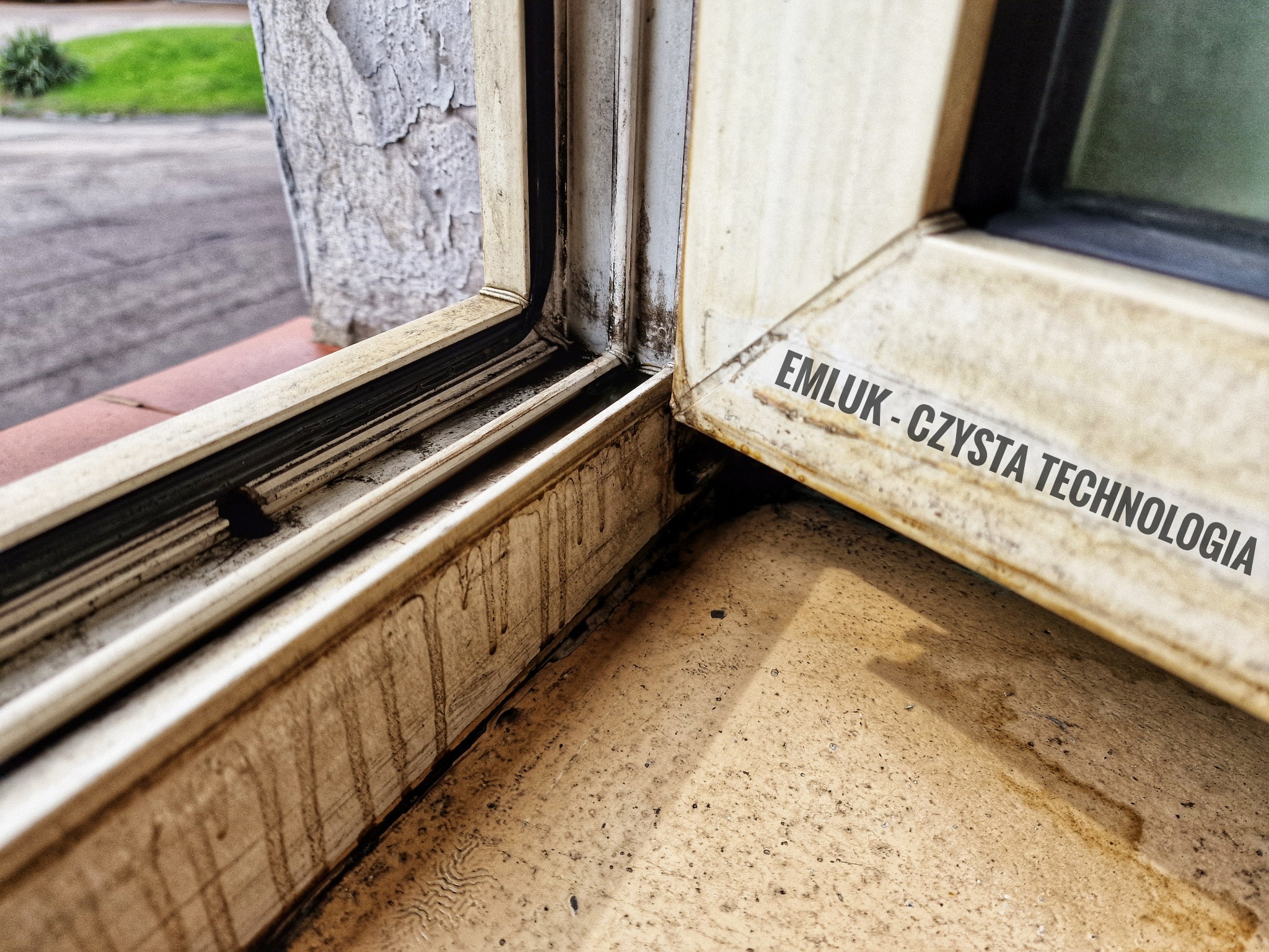Brudne, zaniedbane okno PCV z widocznymi zaciekami i osadami, w rogu ramy widoczny napis 'EMLUK - CZYSTA TECHNOLOGIA'.