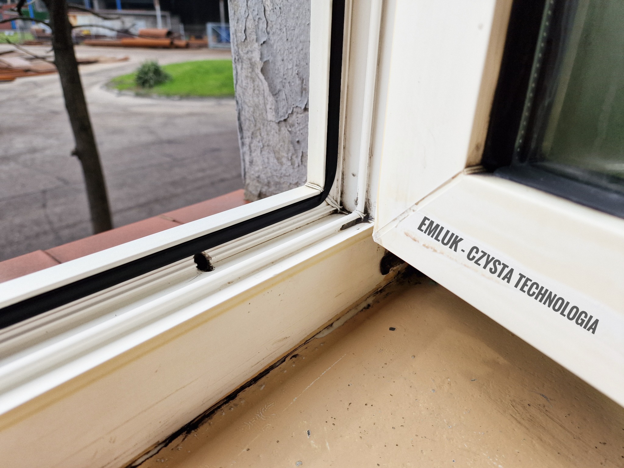 Brudne, nieszczelne okno PCV z widocznym napisem 'EMLUK - CZYSTA TECHNOLOGIA' na ramie, ukazujące zaniedbania i potrzebę czyszczenia lub naprawy.