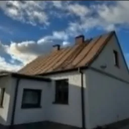 Dom jednorodzinny z dwuspadowym dachem pokrytym brązową dachówką ceramiczną, widoczne kominy i orynnowanie, na tle błękitnego nieba z chmurami.