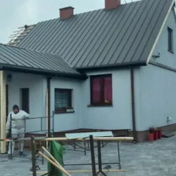 Dom jednorodzinny z szarą blachą na dachu, częściowo zdjęte poszycie dachu, widoczne łaty, robotnicy w trakcie prac, rozstawione rusztowanie, w tle dom z panelami słonecznymi.