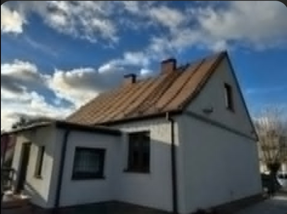 Dom jednorodzinny z dwuspadowym dachem pokrytym brązową dachówką ceramiczną, widoczne kominy i orynnowanie, na tle błękitnego nieba z chmurami.