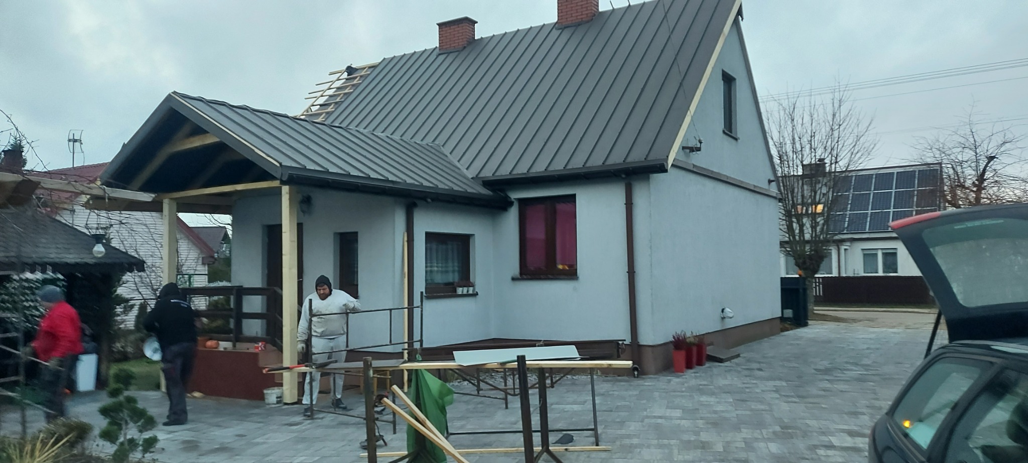 Dom jednorodzinny z szarą blachą na dachu, częściowo zdjęte poszycie dachu, widoczne łaty, robotnicy w trakcie prac, rozstawione rusztowanie, w tle dom z panelami słonecznymi.