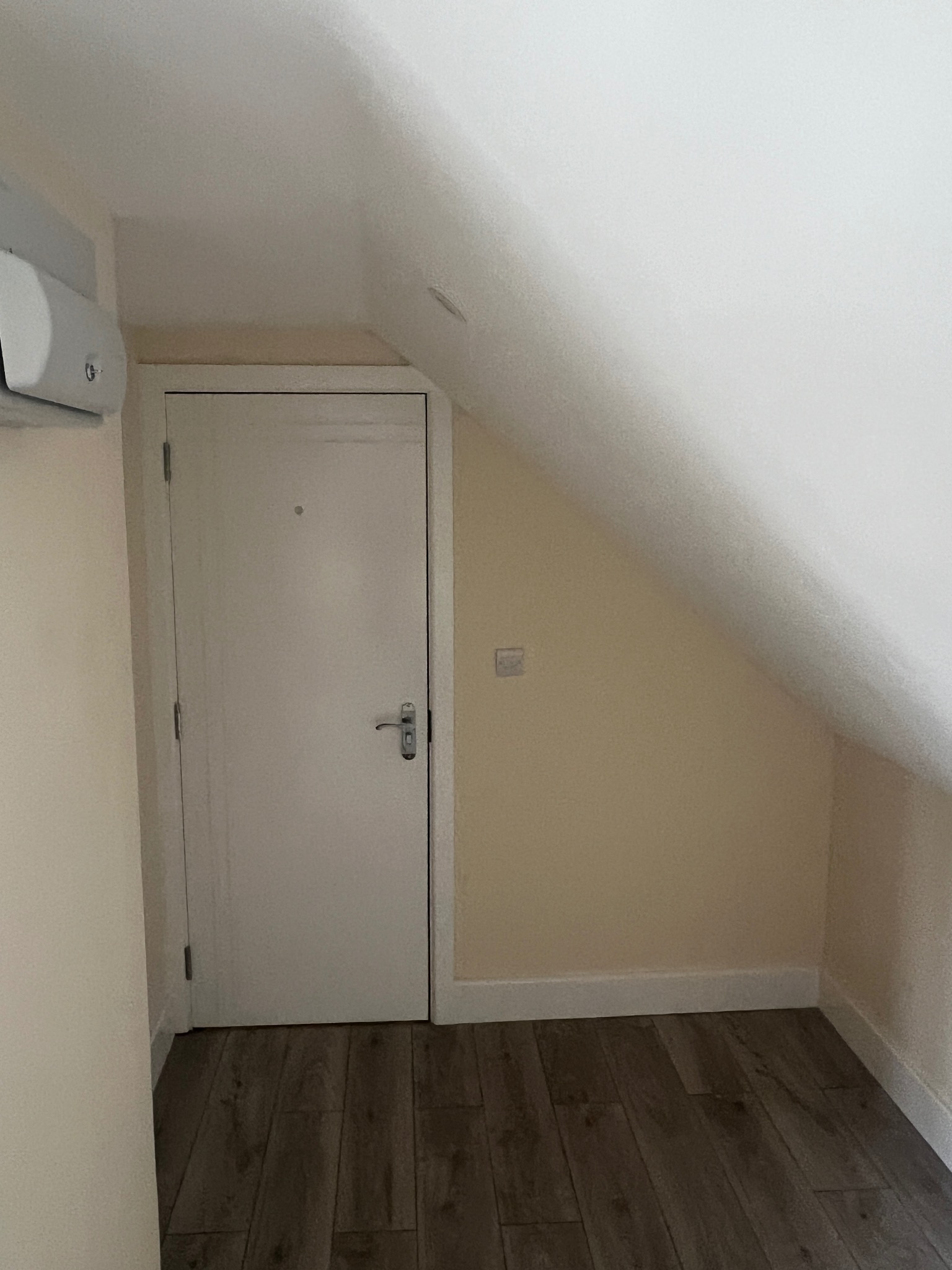 Poddasze z drewnopodobną podłogą, białą ścianą skośną i drzwiami, widoczny klimatyzator na ścianie.