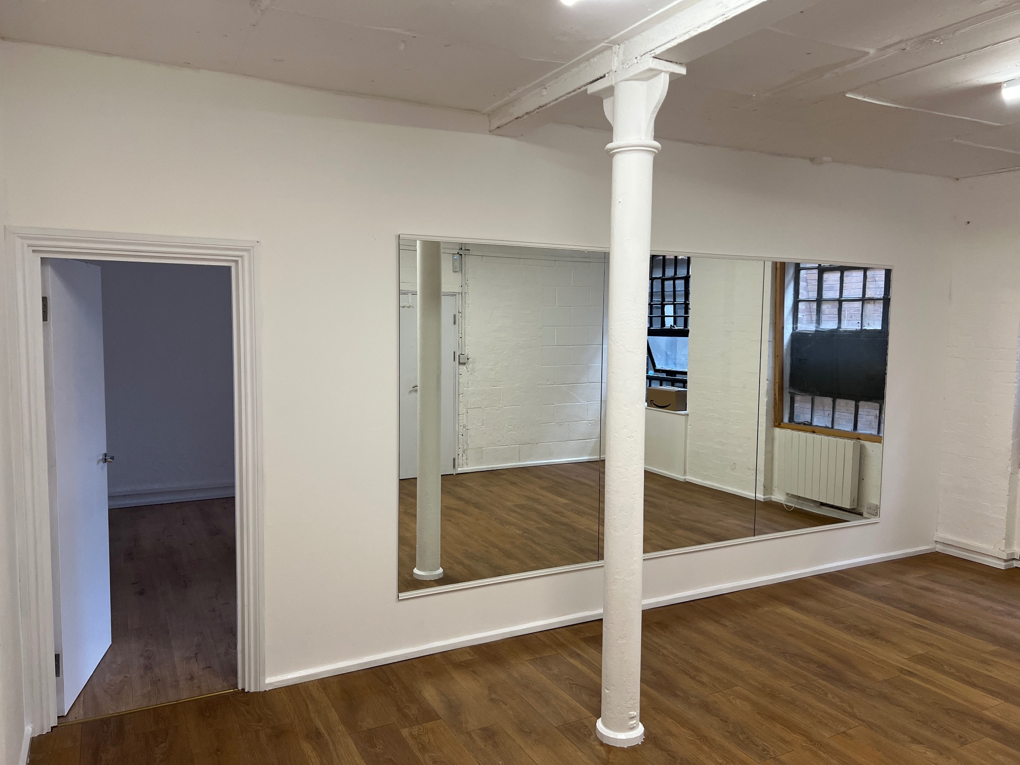 Jasne wnętrze z drewnianą podłogą, duże lustro na ścianie odbija dalszą część pomieszczenia, widoczny biały filar i otwarte drzwi do kolejnego pokoju.