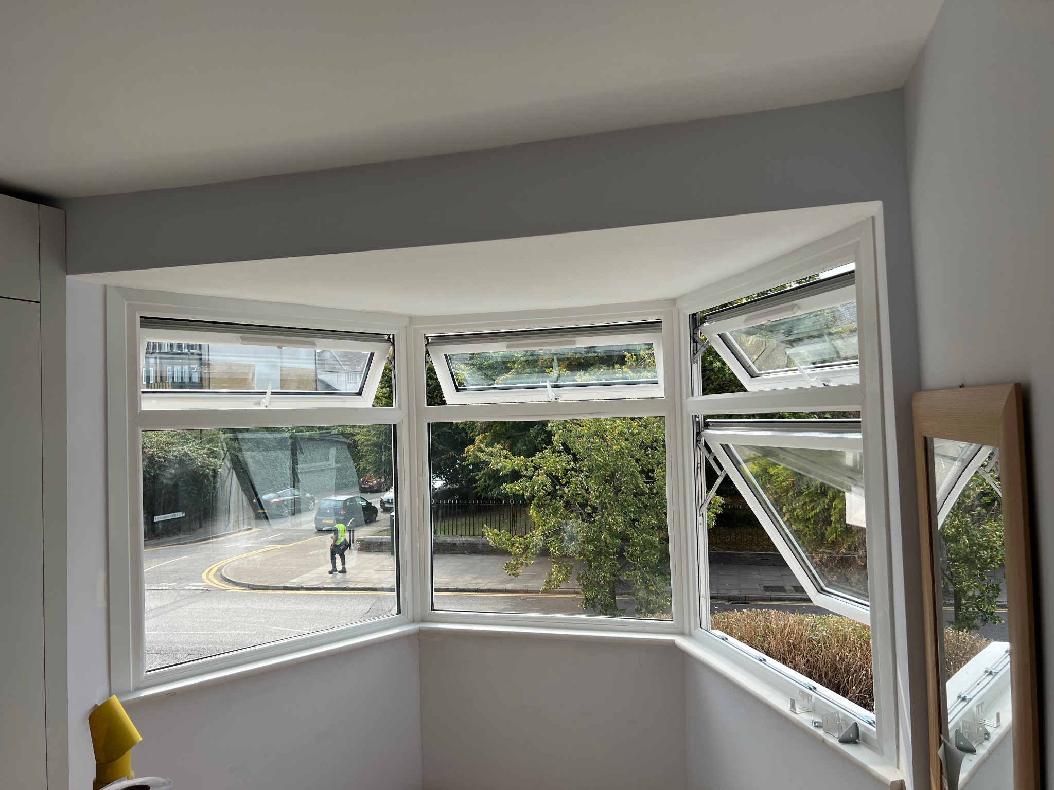 Wnętrze pokoju z oknem wykuszowym, widok na ulicę i chodnik, częściowo otwarte okna uchylne.