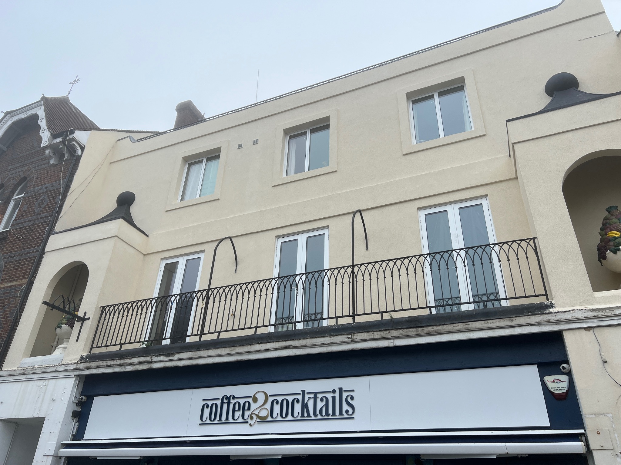 Elewacja budynku po remoncie z beżową farbą, balkon z czarną, kutą balustradą i szyld 'coffee 2 cocktails'.