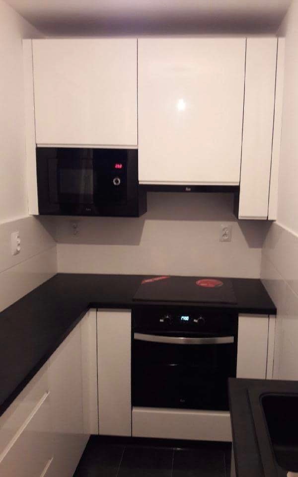 Nowoczesna kuchnia z białymi szafkami, czarnym blatem i sprzętem AGD w zabudowie, widoczne gniazdka elektryczne na ścianie.