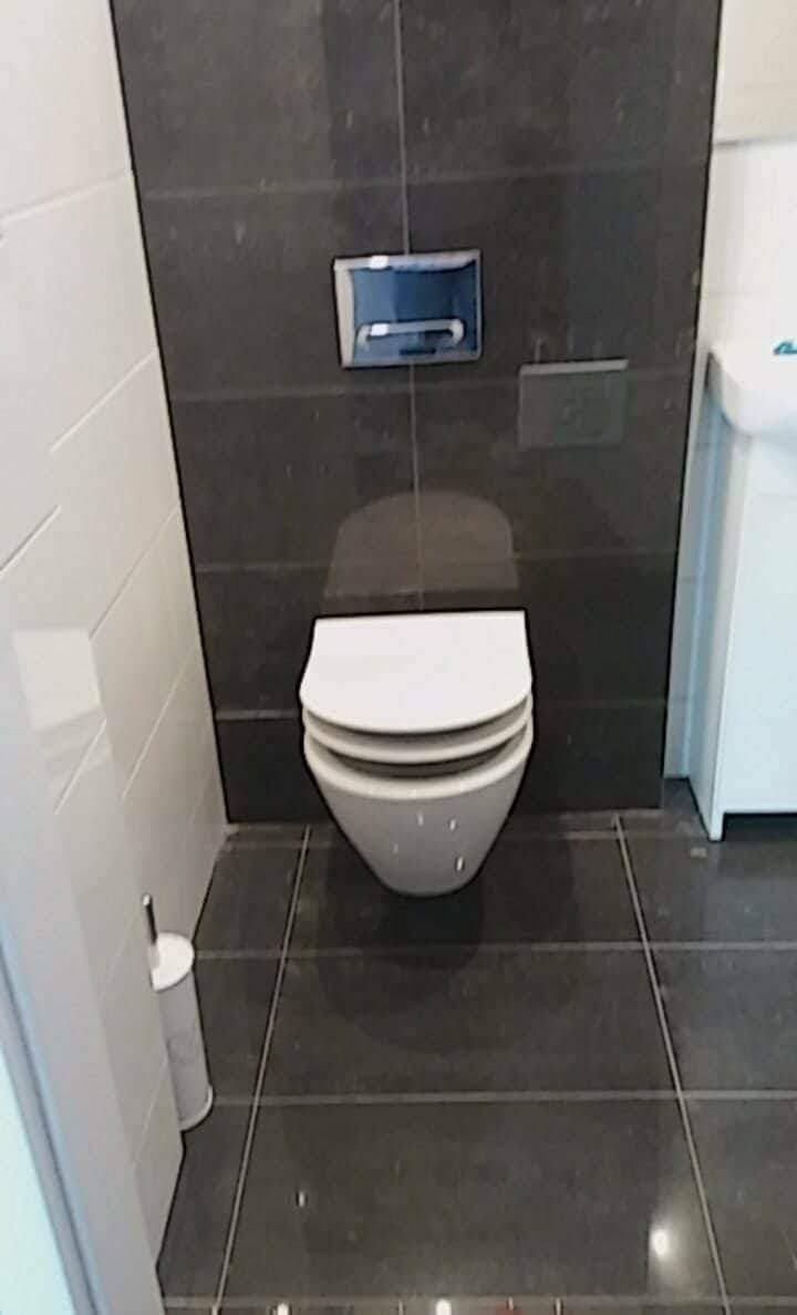 Nowoczesna toaleta podwieszana z chromowanym przyciskiem spłukującym na ciemnej, grafitowej ścianie z płytek, jasne płytki na sąsiedniej ścianie i ciemna podłoga z płytek.