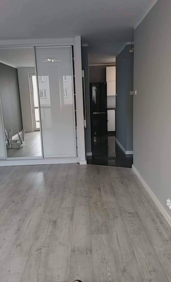 Wykończone wnętrze mieszkania z panelami podłogowymi w odcieniach szarości, widok na szafę z przesuwnymi drzwiami z lustrami oraz aneks kuchenny z czarną lodówką.