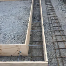 Przygotowanie szalunku i zbrojenia pod fundament, widoczne drewniane deski tworzące formę oraz stalowe pręty zbrojeniowe ułożone wewnątrz, na tle gruzu i żwiru.