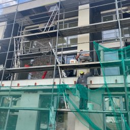 Taurus Bud - Praca na rusztowaniu przy ocieplaniu elewacji budynku, widoczni dwaj pracownicy w trakcie wykonywania prac, jeden nakłada zaprawę z wiadra VASSUD GRUNLIT-SI, w tle ceglana ściana.