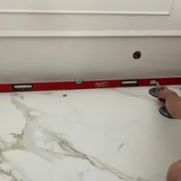 Układanie białych, marmurkowych płytek podłogowych z użyciem poziomicy Milwaukee i przyssawek do przenoszenia płytek. Widoczna dekoracyjna listwa przysufitowa i otwór w ścianie z wystającymi kablami.