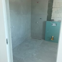 Taurus Bud - Łazienka w trakcie prac wykończeniowych, widoczne szare płytki na ścianach i podłodze, zamontowany stelaż podtynkowy WC z zielonej płyty gipsowo-kartonowej, oraz instalacja wodna.