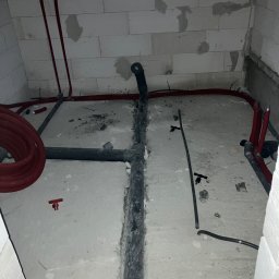 Taurus Bud - Surowa łazienka w trakcie remontu: widoczne czarne rury kanalizacyjne, czerwone rury instalacji wodnej, ściany z betonu komórkowego z widocznymi śladami po łatach, betonowa podłoga.