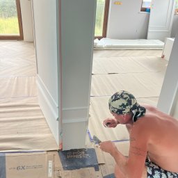 Taurus Bud - Mężczyzna w kamuflażowej chuście szpachluje listwę przypodłogową przy drzwiach w nowym domu, na podłodze kartony zabezpieczające z nadrukiem '6X More Silent than Rigid Core Laminate'.