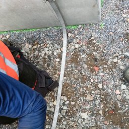 Taurus Bud - Mocowanie metalowej listwy do betonowej konstrukcji na zewnątrz, widoczne kamienie i żwir, fragment ubrania roboczego.