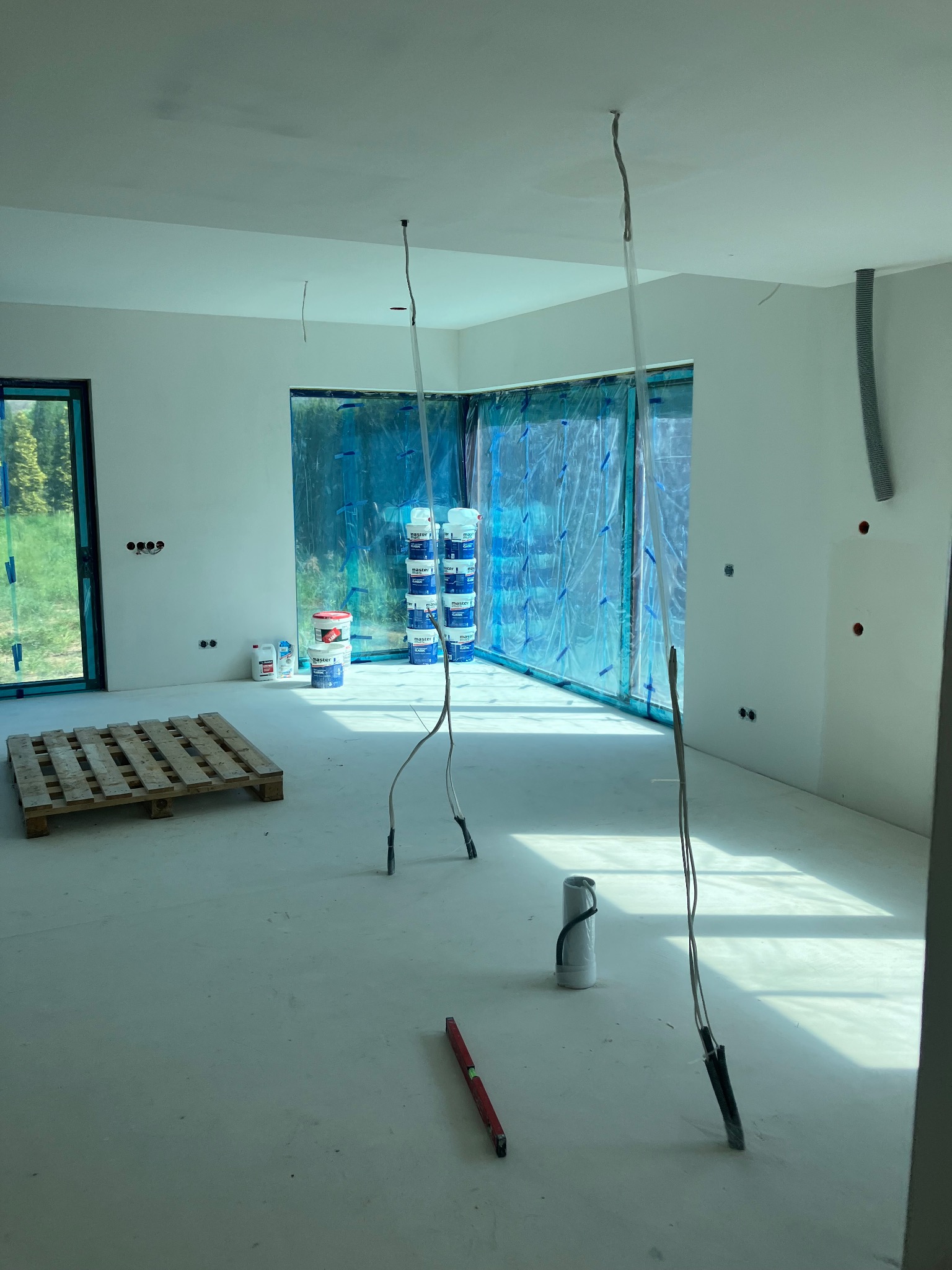 Wnętrze w trakcie przygotowań do prac malarskich: zabezpieczone okna folią, puszki z farbą, poziomica na podłodze, przygotowane przewody elektryczne.