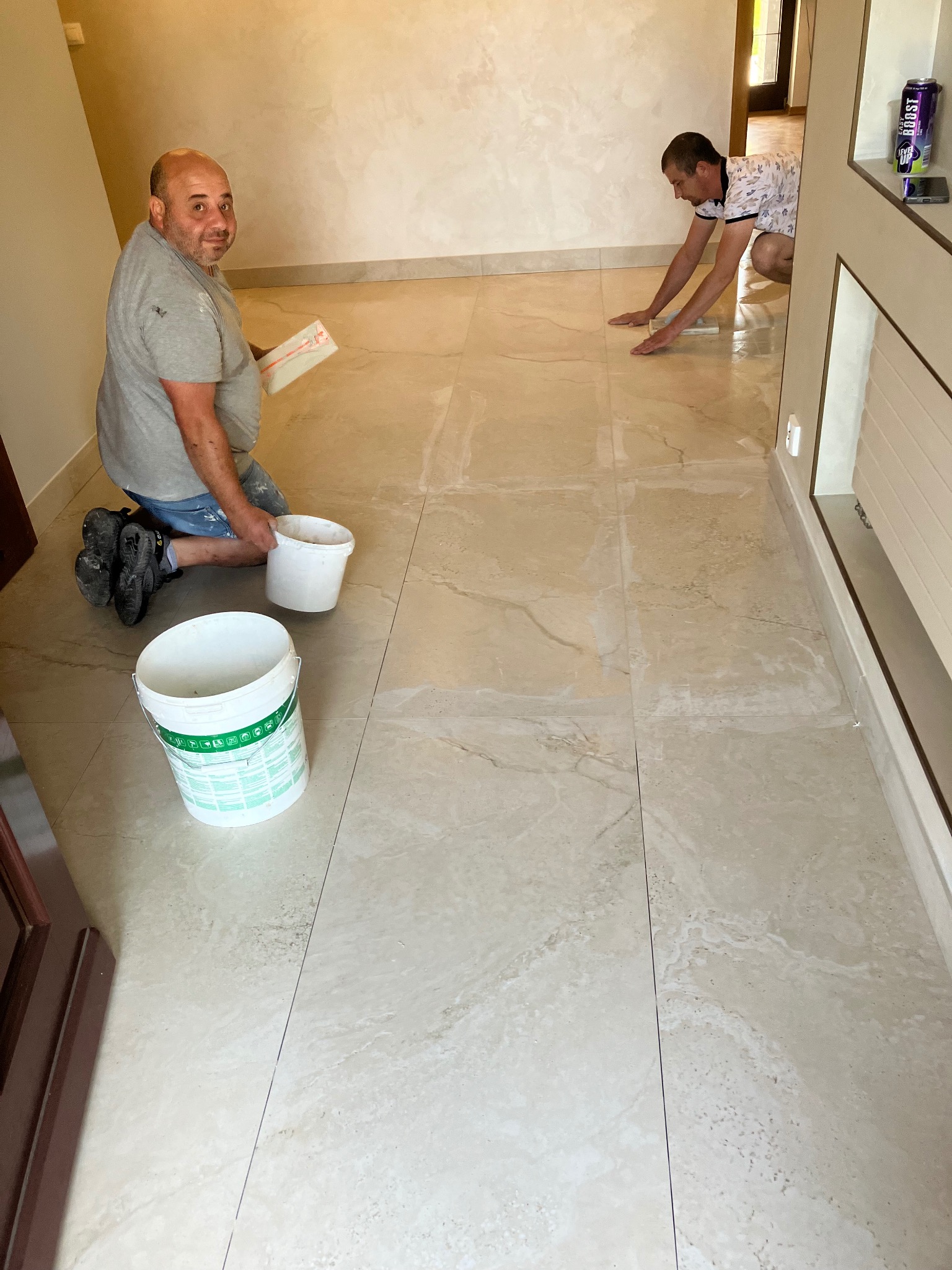 Dwóch mężczyzn podczas układania beżowych, marmurkowych płytek podłogowych w korytarzu, widoczne wiadra z zaprawą i narzędzia.