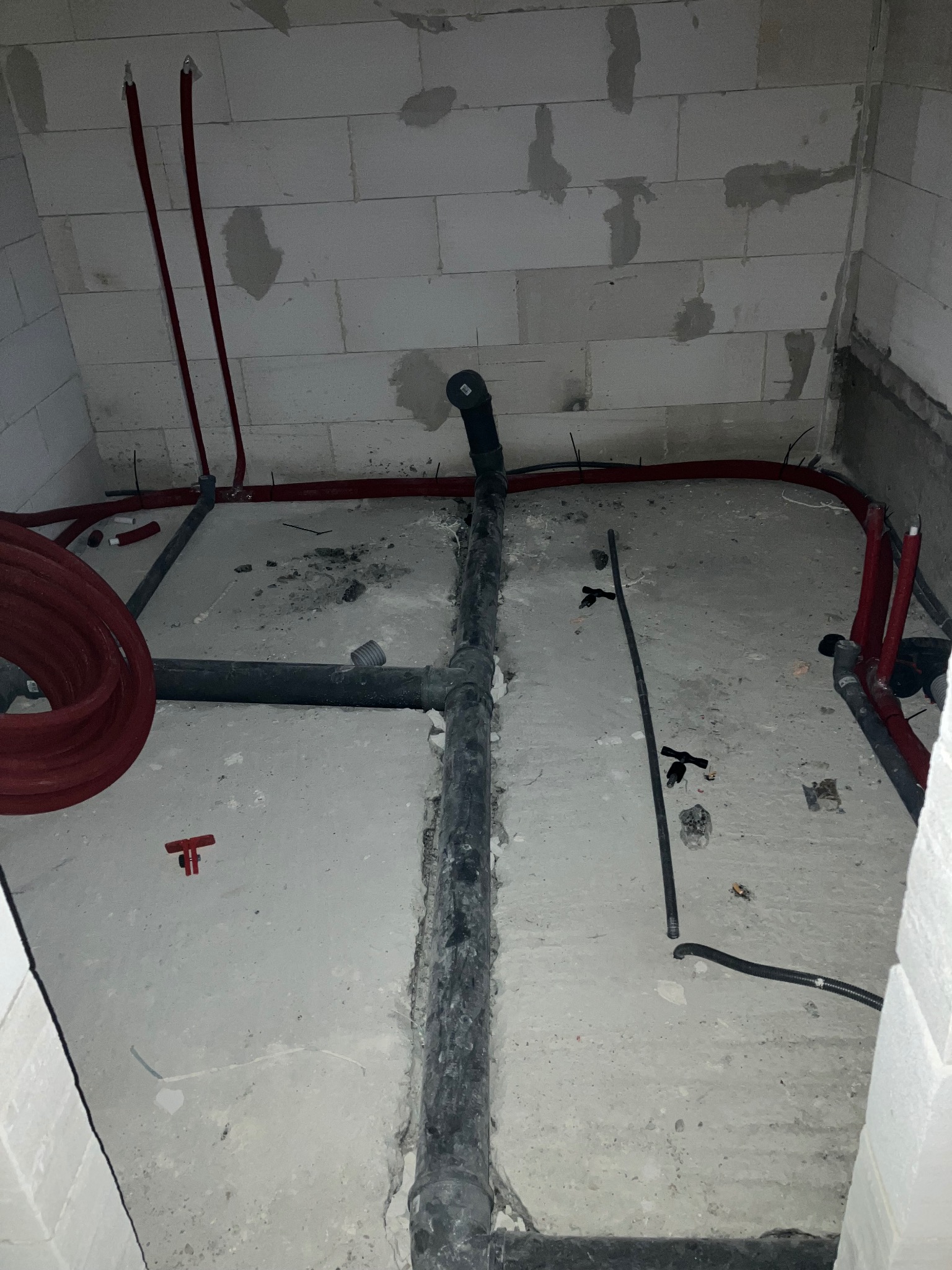 Surowa łazienka w trakcie remontu: widoczne czarne rury kanalizacyjne, czerwone rury instalacji wodnej, ściany z betonu komórkowego z widocznymi śladami po łatach, betonowa podłoga.