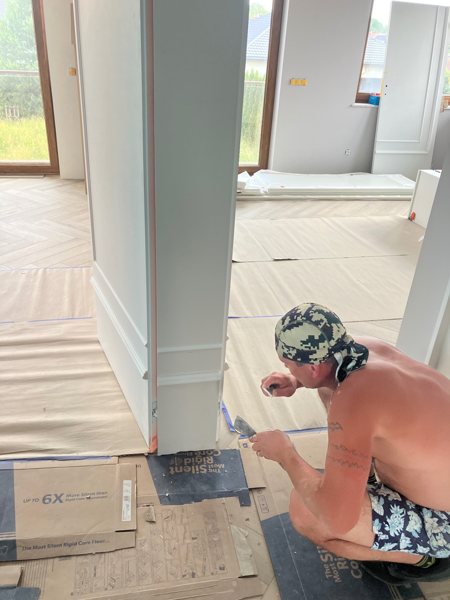 Mężczyzna w kamuflażowej chuście szpachluje listwę przypodłogową przy drzwiach w nowym domu, na podłodze kartony zabezpieczające z nadrukiem '6X More Silent than Rigid Core Laminate'.