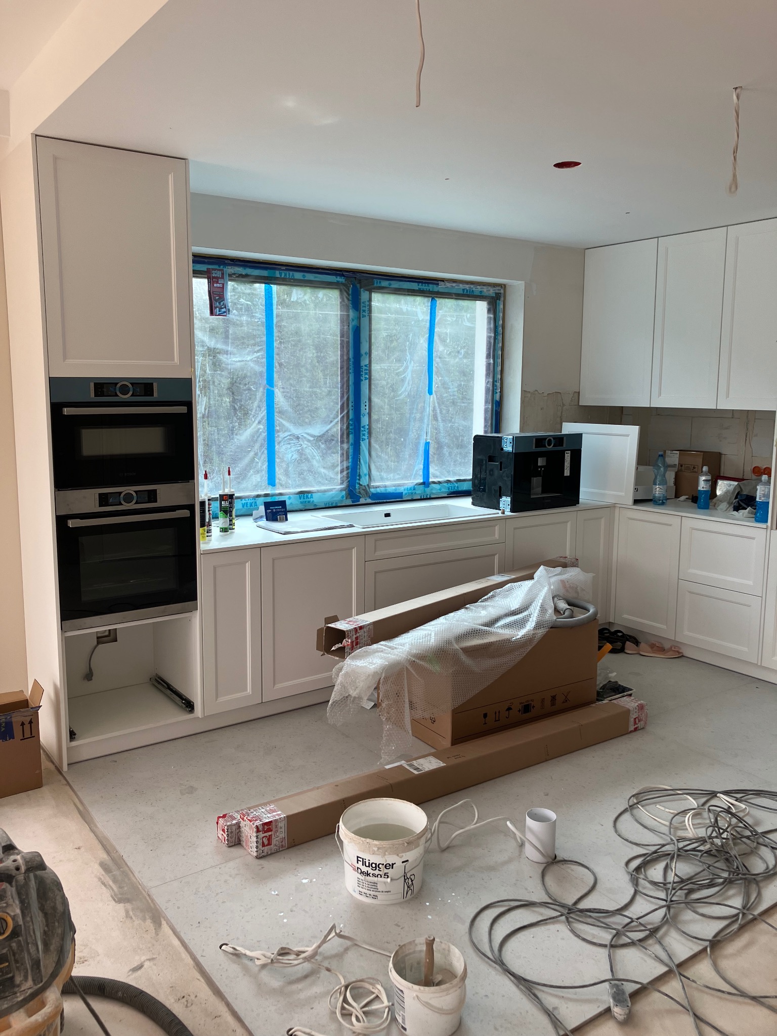 Kuchnia w trakcie montażu: białe szafki kuchenne z wbudowanym piekarnikiem i mikrofalówką, okno zabezpieczone folią, na podłodze materiały budowlane, kartony i wiadro z farbą Flugger Dekso 5.