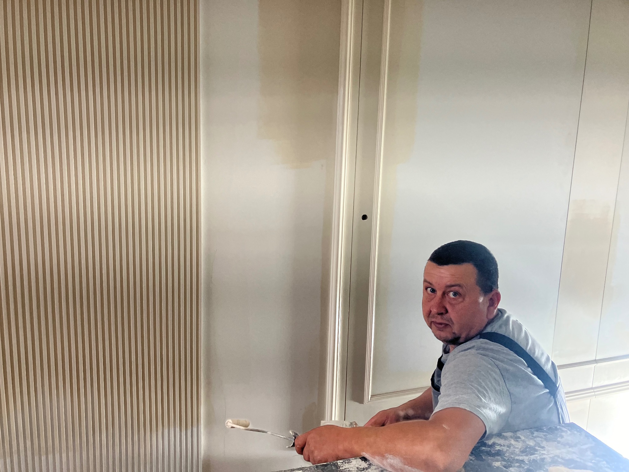 Mężczyzna maluje ścianę wałkiem, widoczne pionowe panele dekoracyjne i fragment pomalowanej ściany z ozdobną listwą.