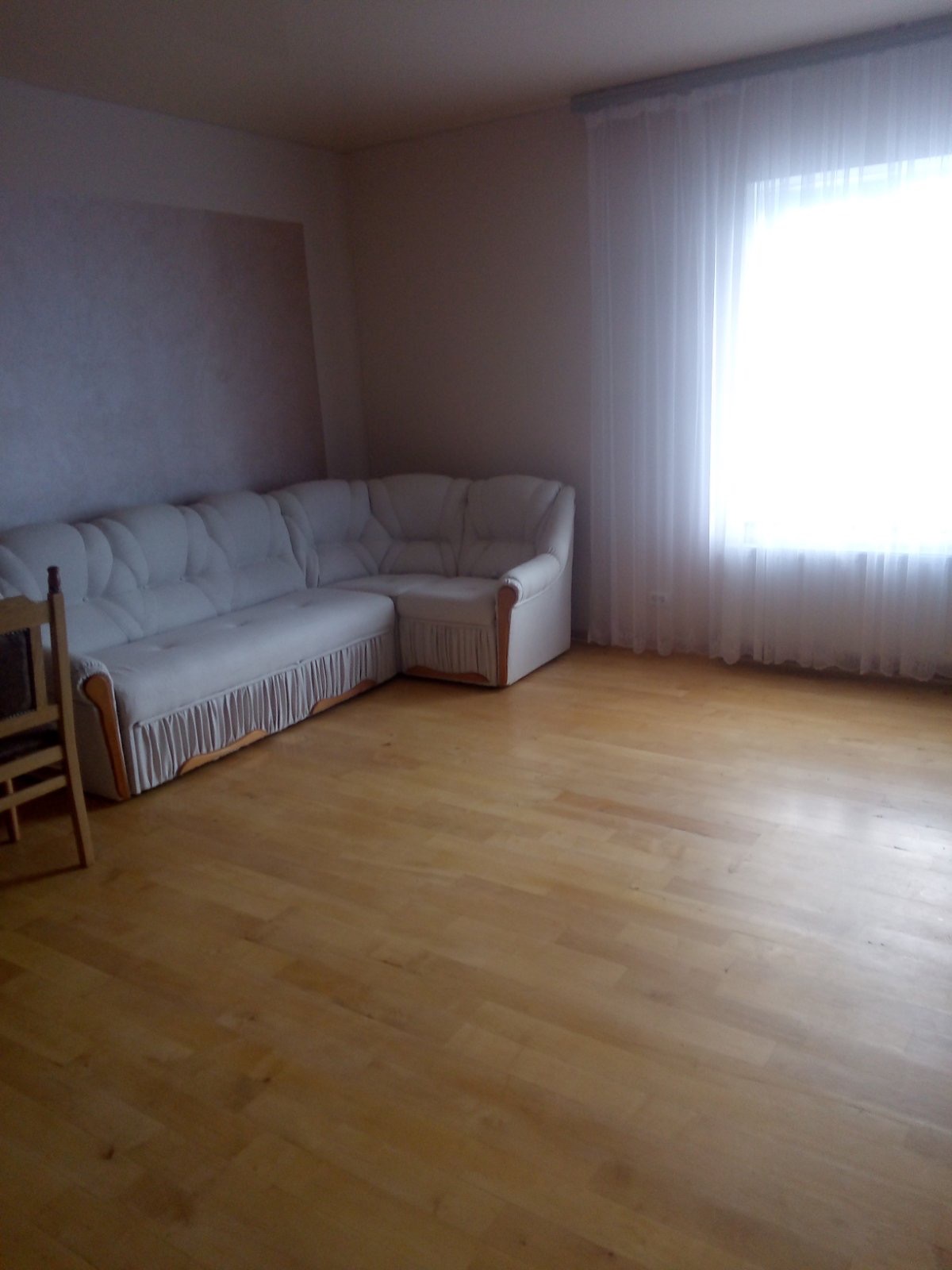 Pusty pokój z drewnianą podłogą, białym narożnikiem z falbanką i jasnymi zasłonami w oknie.