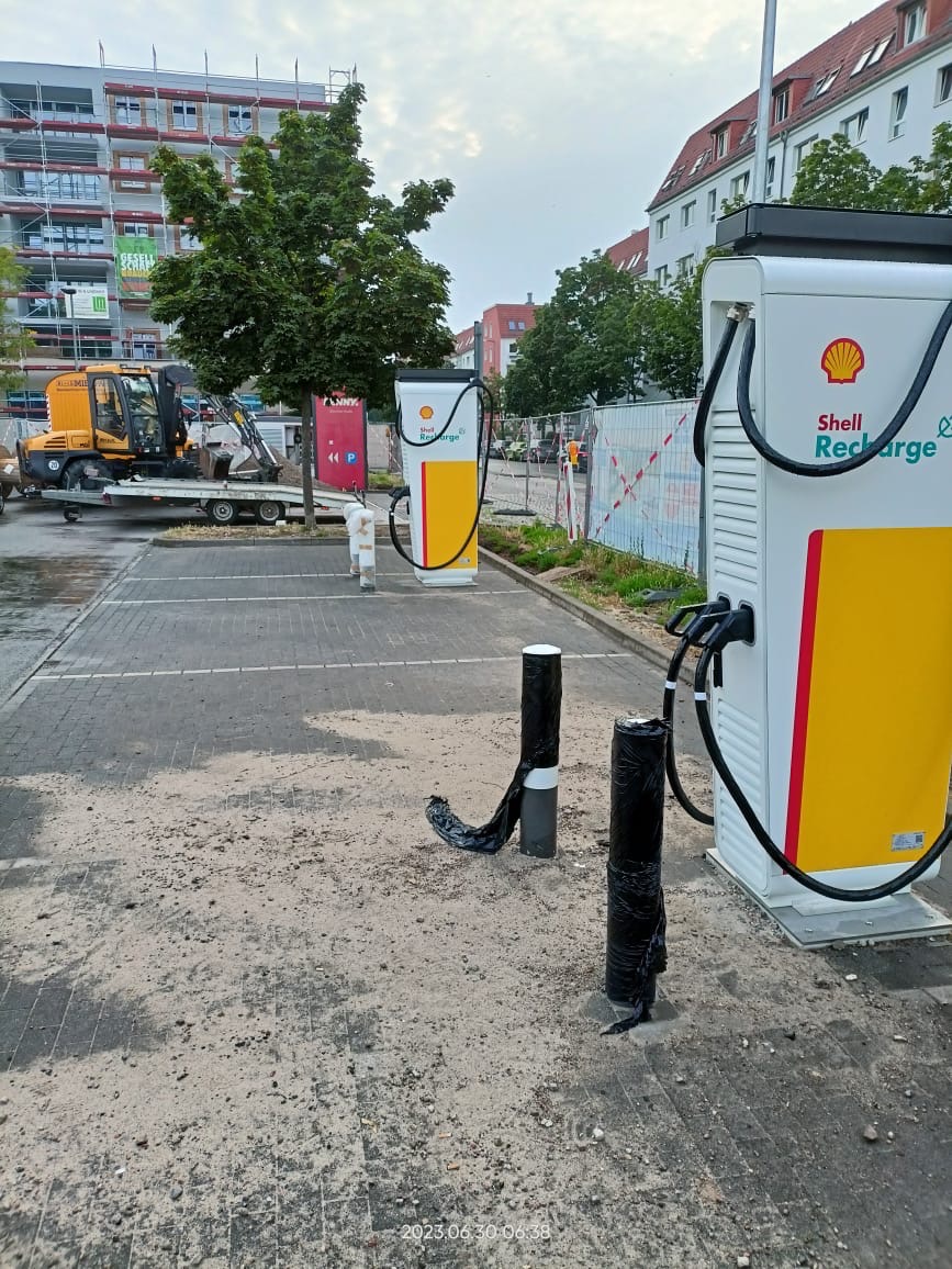 Dwie stacje ładowania Shell Recharge na parkingu z widocznymi śladami piasku i budowy, w tle budynek w rusztowaniach i mała koparka na przyczepie.