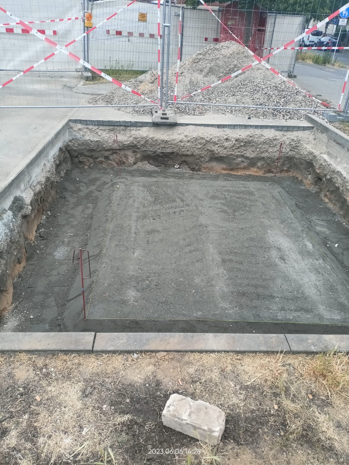 Wybetonowany wykop pod fundamenty z widocznymi stalowymi prętami i sznurkami wyznaczającymi granice, zabezpieczony siatką i taśmą ostrzegawczą, obok sterta gruzu.