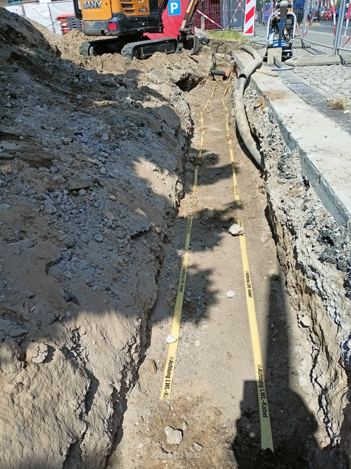 Wykop w ziemi z widocznymi żółtymi taśmami ostrzegawczymi 'Achtung LWL-Kabel', mała koparka SANY i sprzęt budowlany w tle.