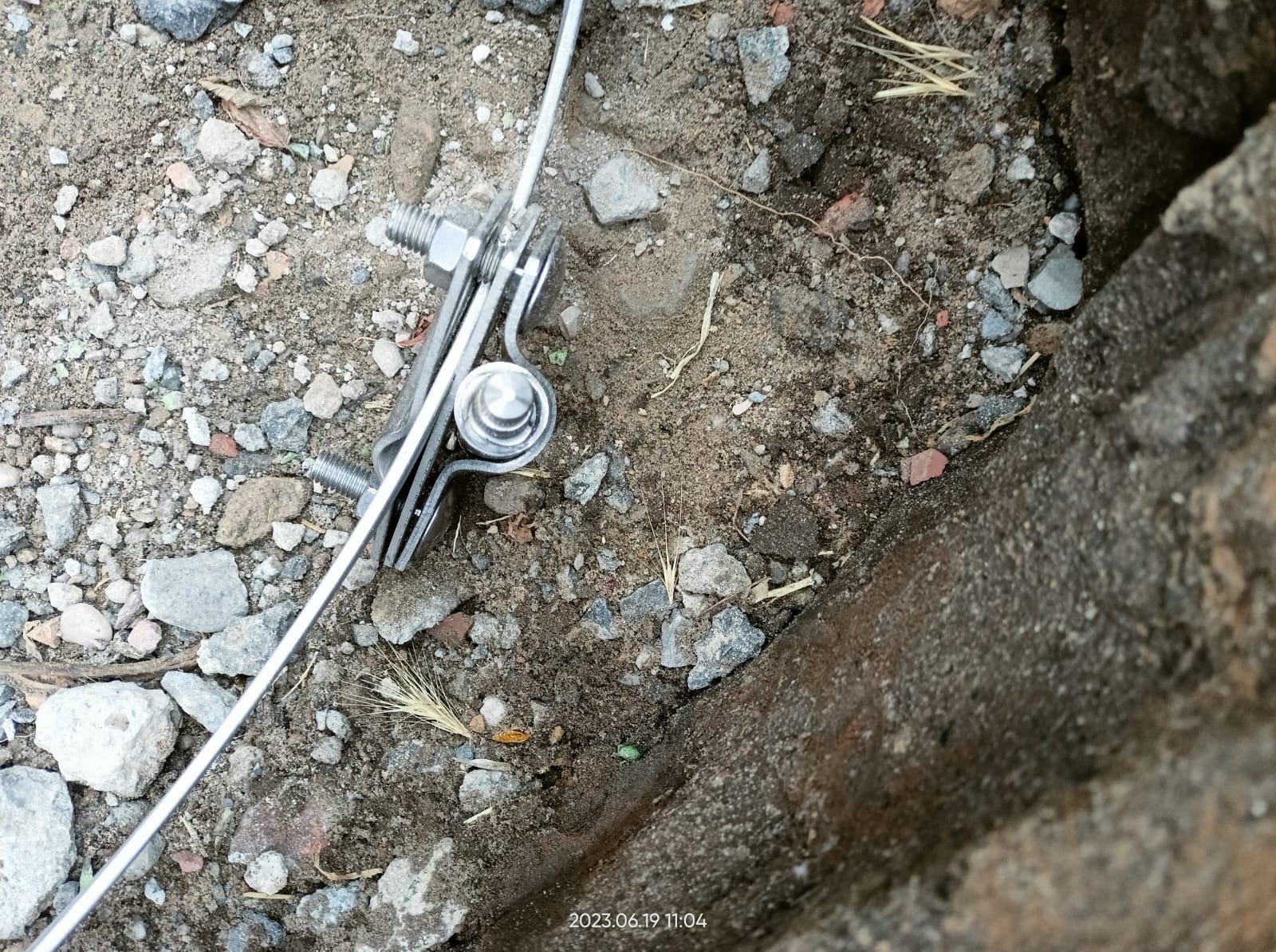 Mocowanie przewodu odgromowego do betonowej podmurówki na tle ziemi z kamieniami.