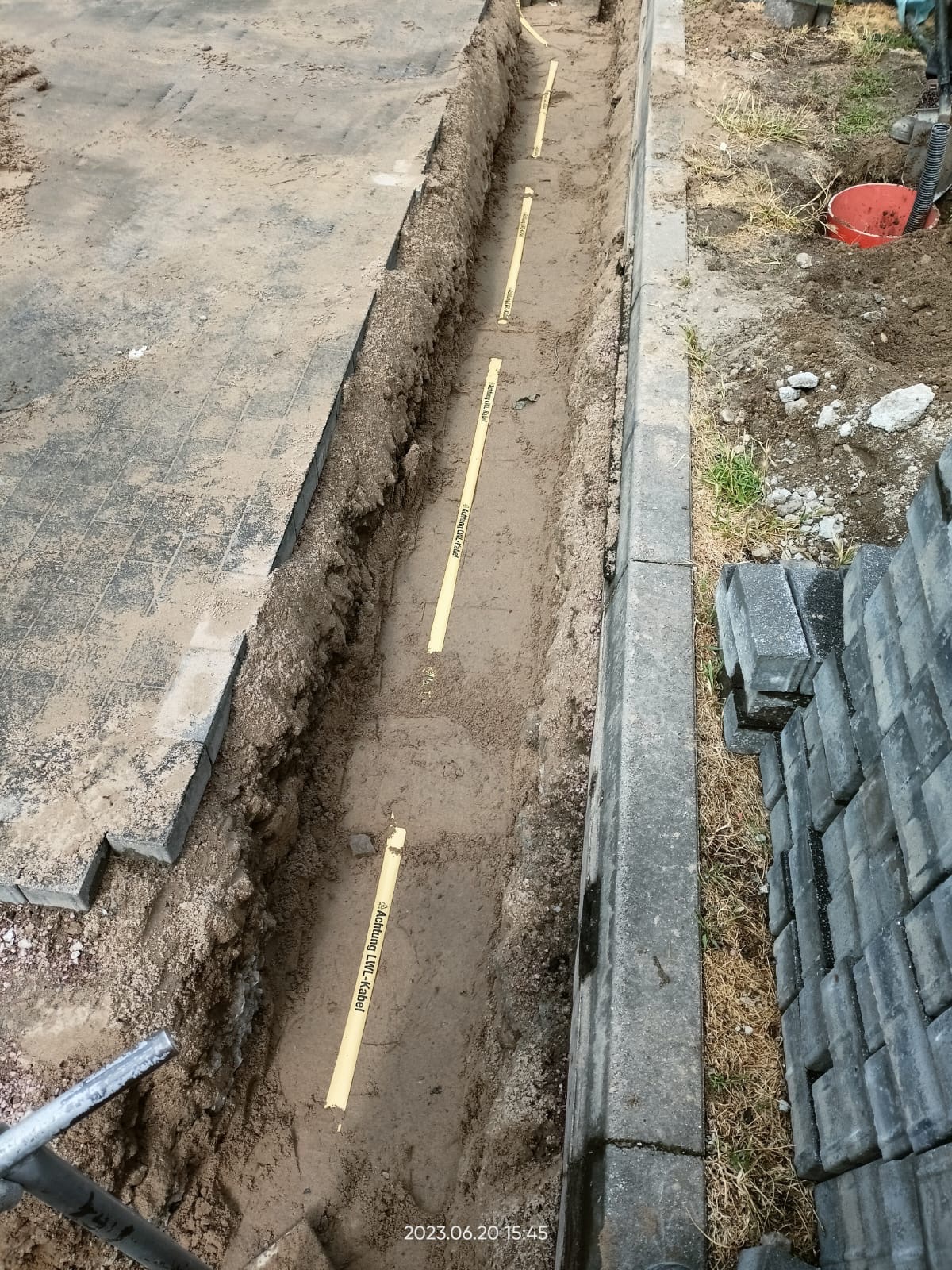 Wykop pod kable z ułożonymi rurami ochronnymi 'Achtung LWL-Kabel' wzdłuż krawężnika i kostki brukowej, widoczne narzędzia i wiadro.