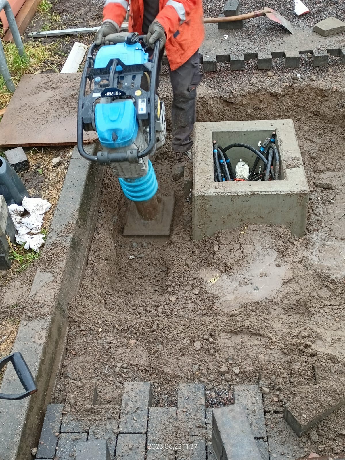 Pracownik w pomarańczowej kurtce zagęszcza grunt ubijarką wibracyjną obok betonowej studzienki z kablami i rurami, przygotowując podłoże pod ułożenie kostki brukowej.